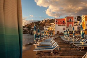 A beach with rows of sun loungers and umbrellas, a hillside with colorful buildings, and a clear sky.