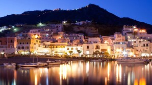A coastal town at dusk with buildings along the waterfront, boats docked, and a hill in the background.