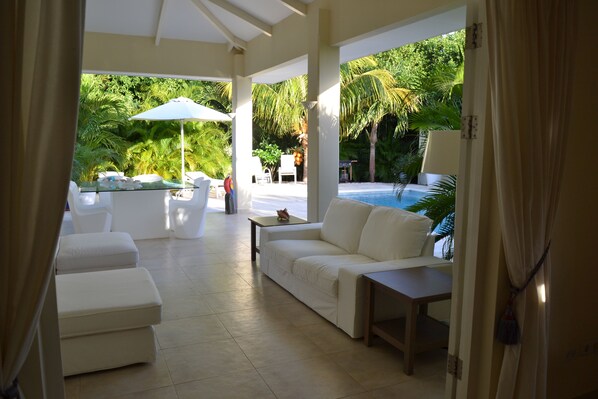 A spacious indoor area with a white sofa, a small table, and a glass partition. There is a pool visible through the glass, surrounded by lush greenery.