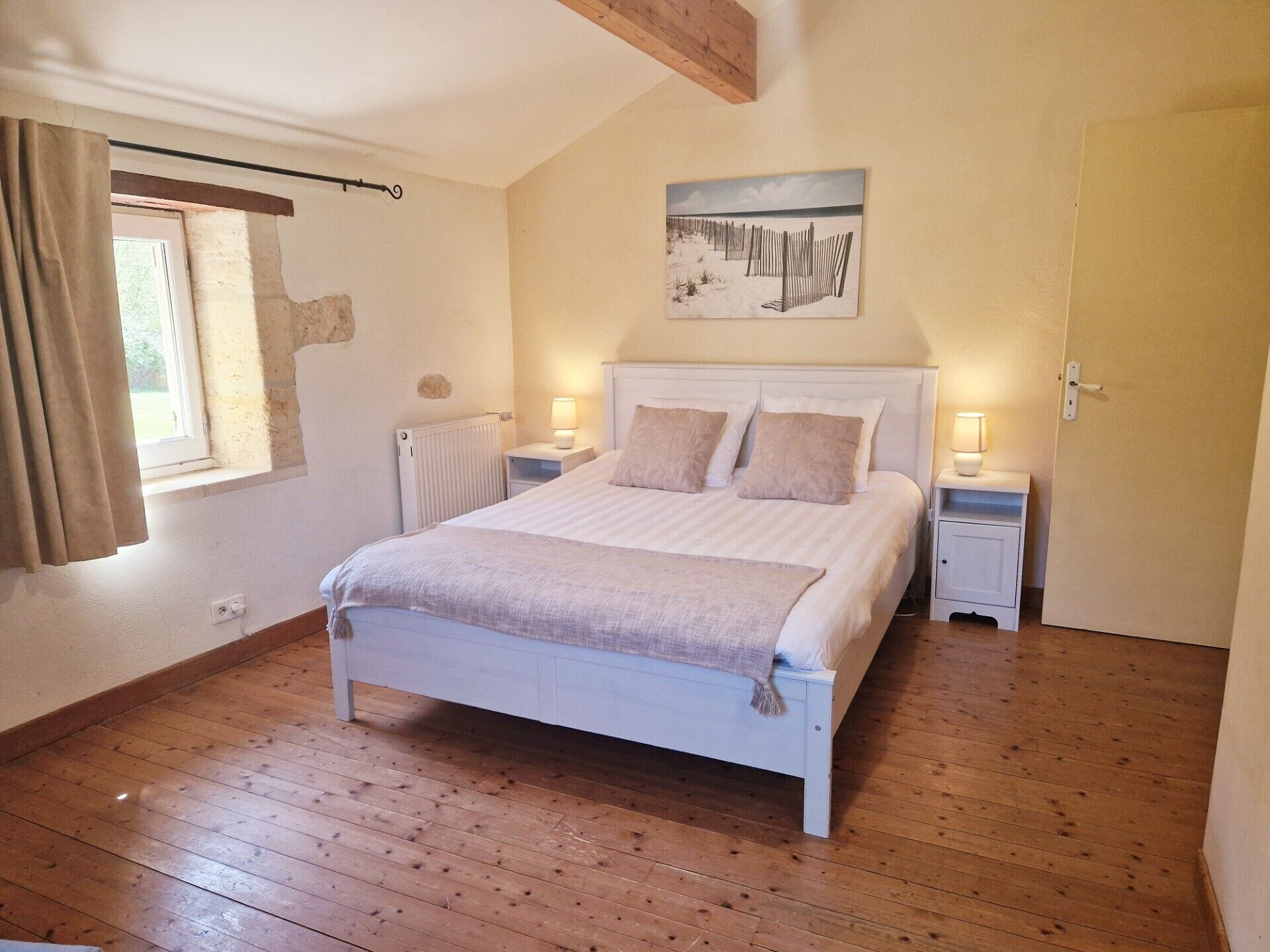 Une chambre avec un plancher en bois, un lit doté d’une tête de lit blanche, deux tables de chevet avec des lampes, une fenêtre avec des rideaux et un tableau accroché au mur.