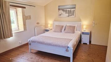 Une chambre avec un plancher en bois, un lit doté d’une tête de lit blanche, deux tables de chevet avec des lampes, une fenêtre avec des rideaux et un tableau accroché au mur.