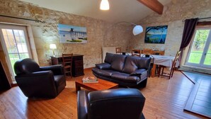 A spacious living room with wooden flooring, a black leather sofa, a coffee table, and a dining area with a table and chairs. - Le Moulin charming cottage near St Emilion (Lugaignac)