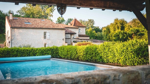 A pool area with a stone wall, a white building with a tiled roof, and lush greenery. - Le Moulin charming cottage near St Emilion (Lugaignac)
