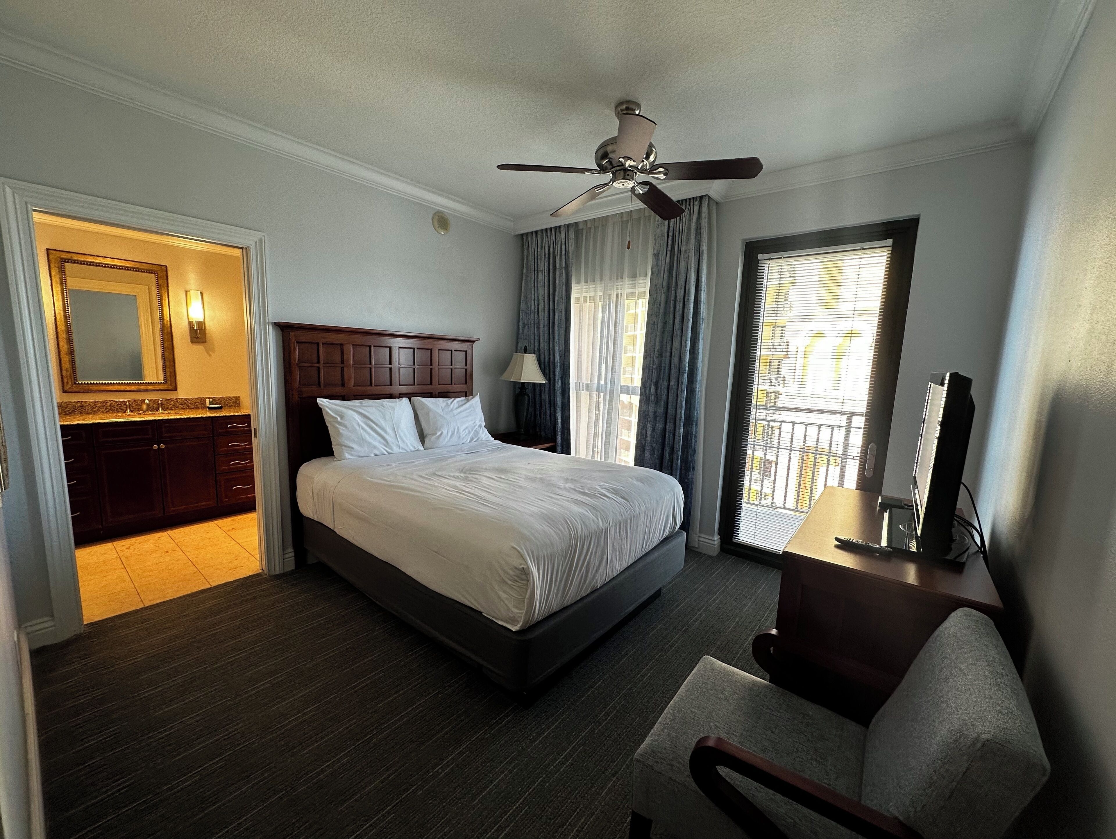 A hotel room with a bed, a desk with a computer, a chair, a ceiling fan, a window with curtains, and a bathroom with a mirror and vanity.