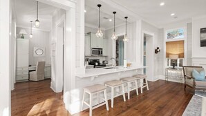 A modern kitchen with white cabinetry, a central island with stools, pendant lighting, and a view of the dining area. - Luxury Living Along 30A-6 Seater Golf Cart/6 bikes! Next to Waterpark & Beach (Santa Rosa Beach)