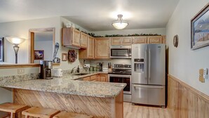 A kitchen with wooden cabinets, granite countertops, a stainless steel refrigerator, a microwave, and a stove. There are two wooden stools and a light fixture above the counter. - Beautiful Condo, Sleeps 8, Gorgeous Views, Wood Fireplace, HDTV, Hot Tubs - BB (Silverthorne)