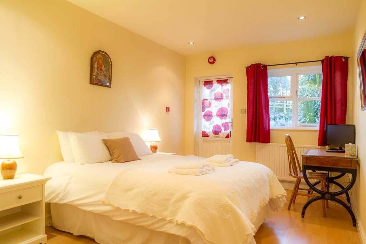 A bedroom with a bed, bedside tables, a desk, a chair, a window with curtains, and a wall-mounted television.