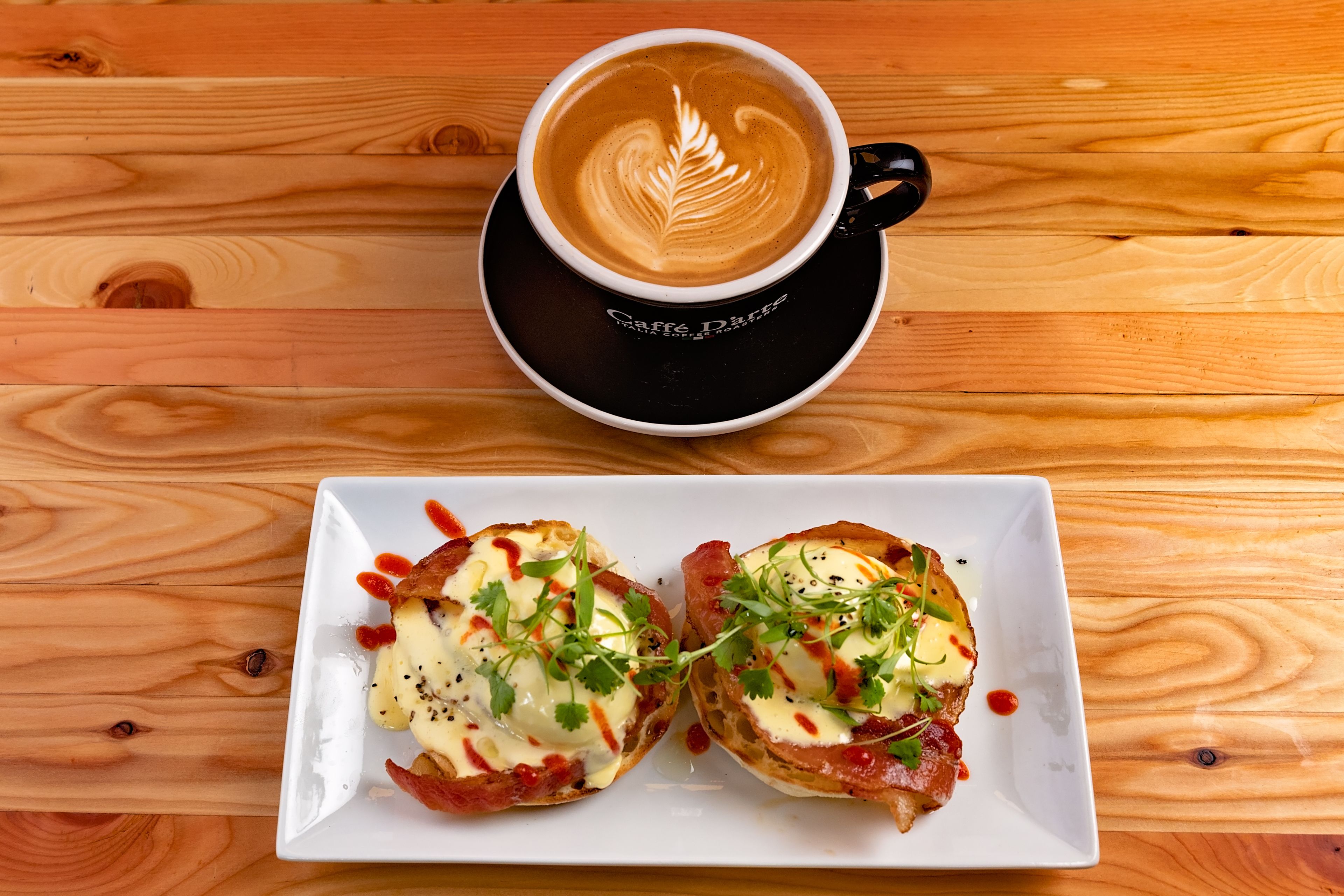 A wooden table with a black cup and saucer labeled "Caffe D'Angelo" and a white plate with two open-faced sandwiches topped with cheese, herbs, and sauce.