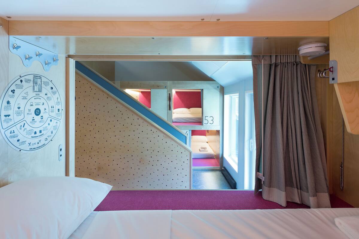 A hotel room with a bunk bed, a wooden wall with a circular diagram, a staircase, and a window with curtains.