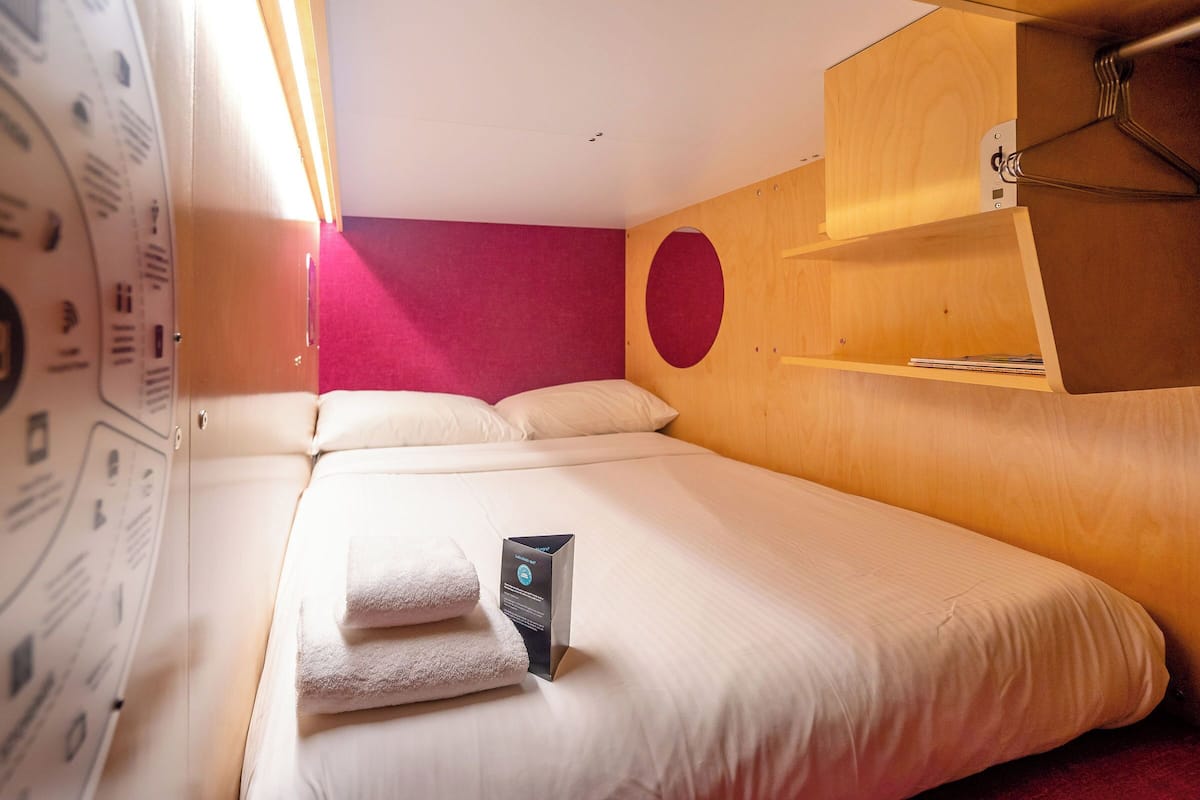A neatly made bed with white linens and a black object on it, a wooden shelf above the bed, and a red wall behind the bed.