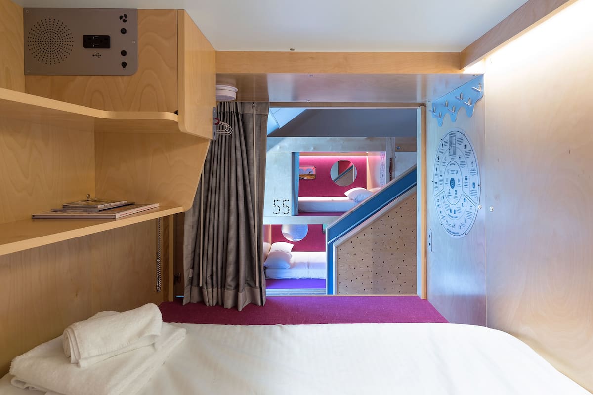 A compact room with a bunk bed, a wooden shelf with books, a curtain, and a wall-mounted control panel.