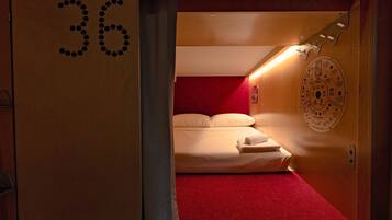 Une chambre d’hôtel avec un tapis rouge, un lit recouvert de draps blancs, une porte en bois portant le numéro 36 et une horloge murale.