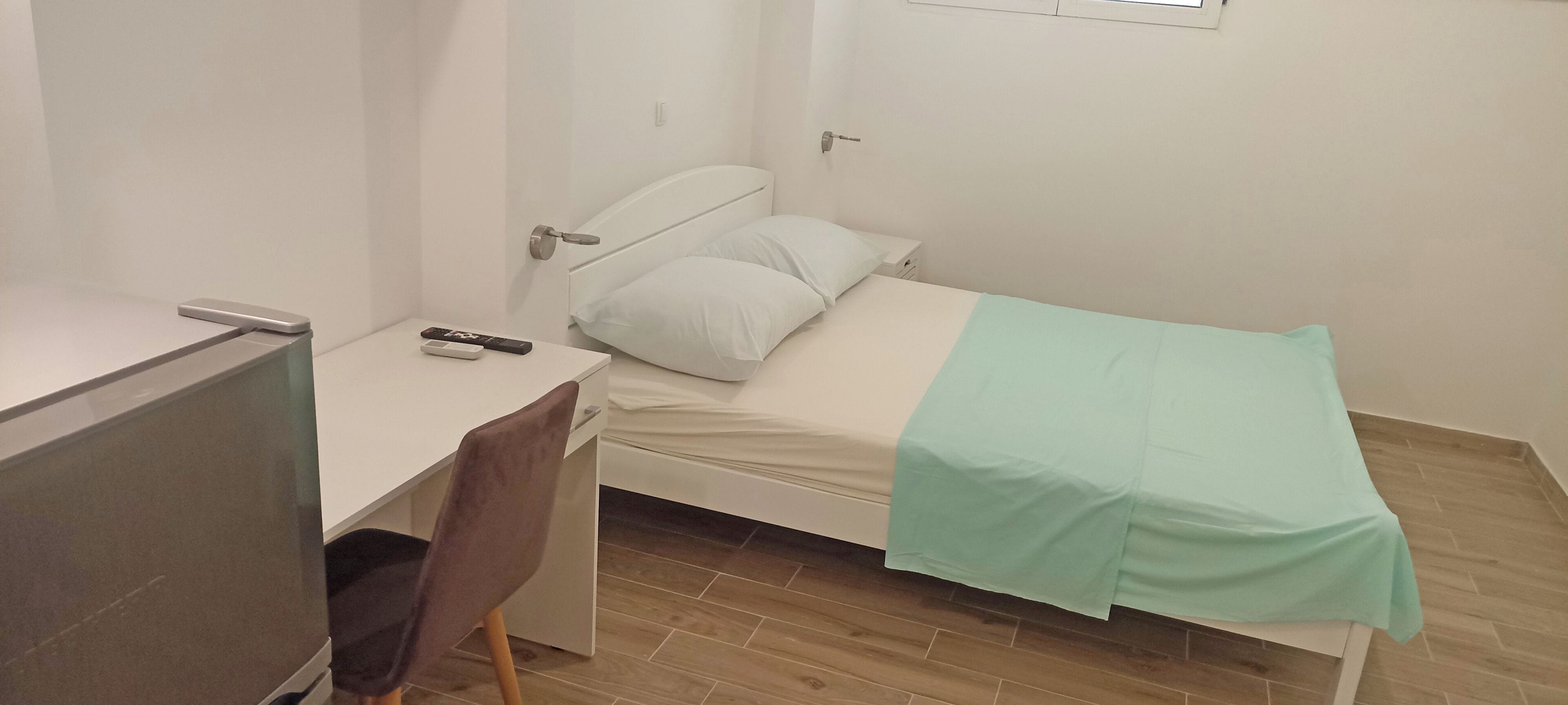 Una habitación pequeña y limpia con una cama, un escritorio y una silla. Hay un control remoto sobre el escritorio.
