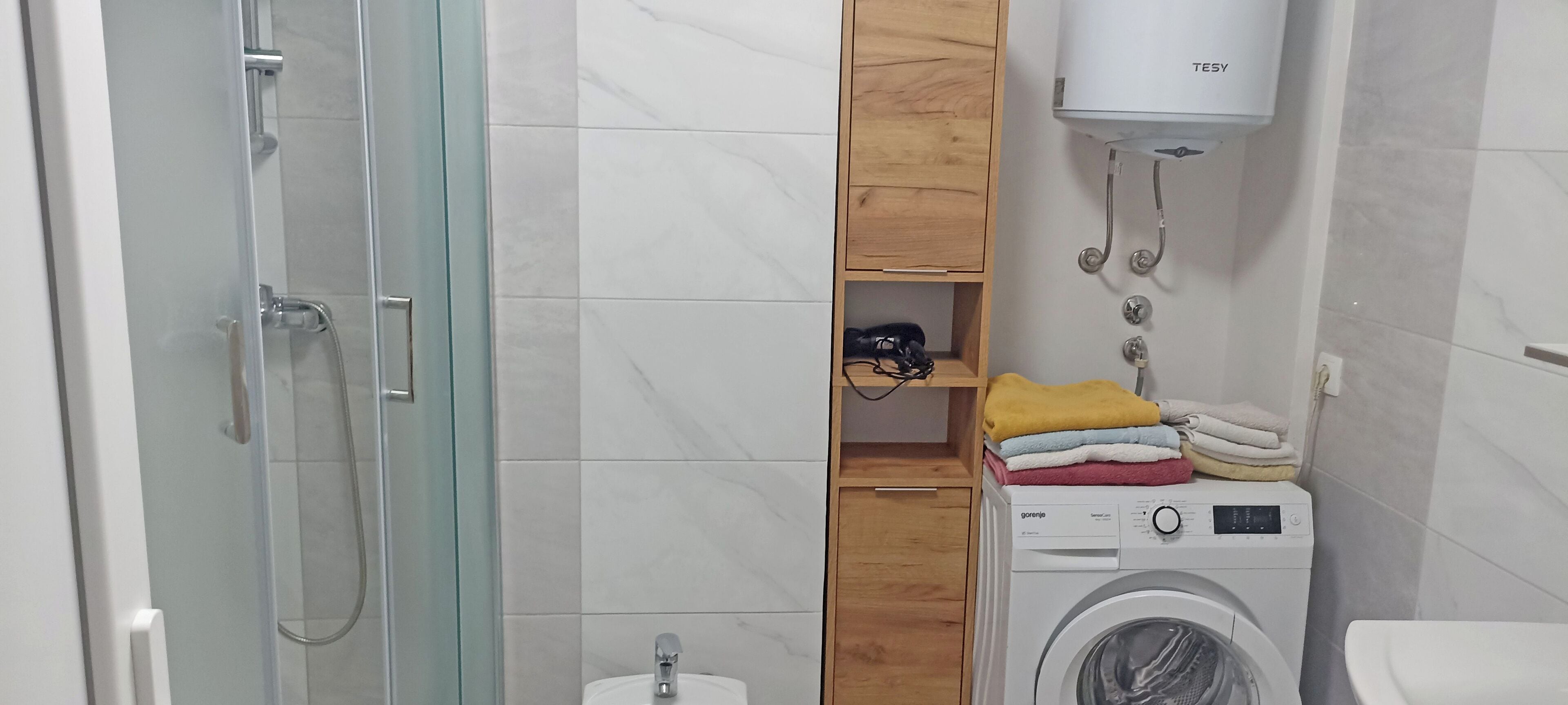A bathroom with a glass shower enclosure, a wooden shelf with folded towels, a washing machine, and a toilet.