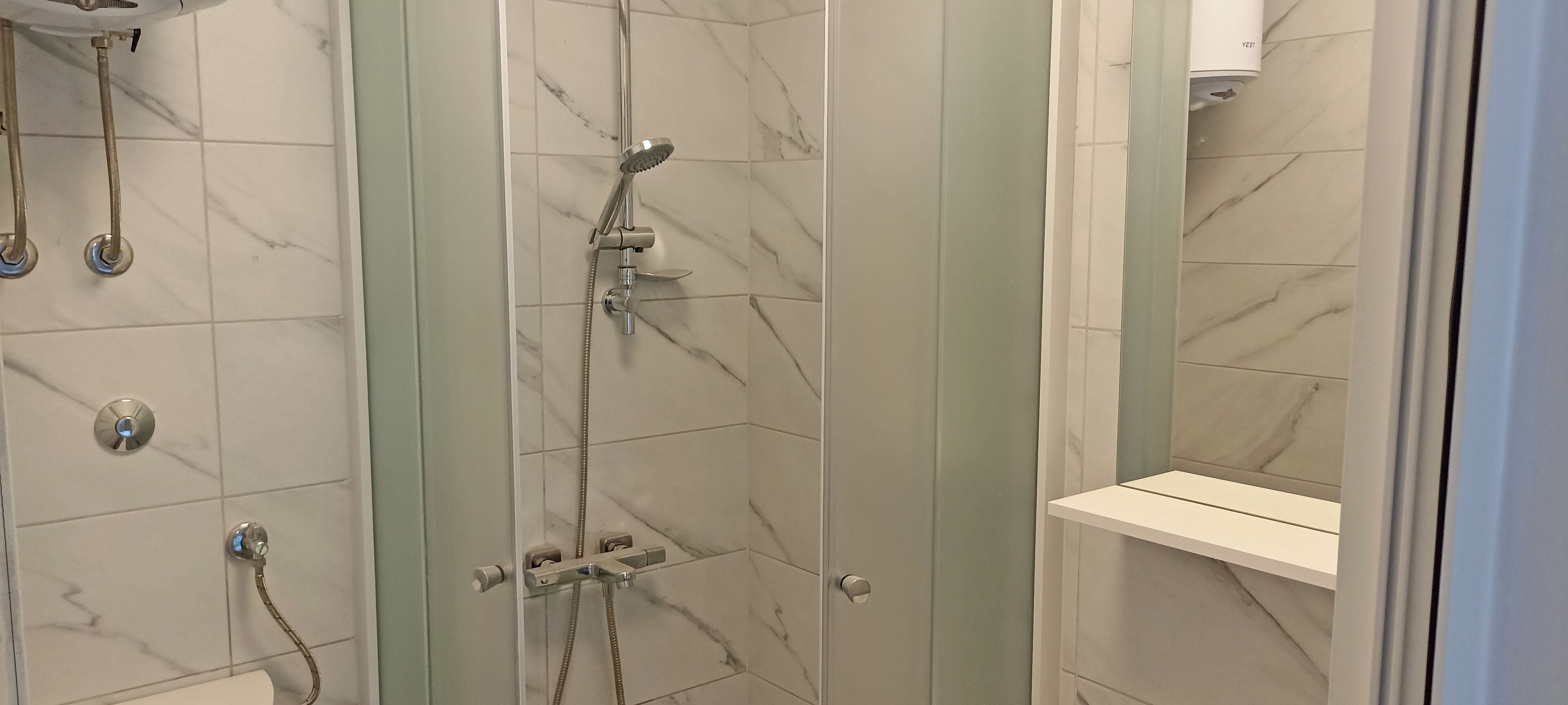 A bathroom with a glass-enclosed shower, marble tiles, and a white shelf.