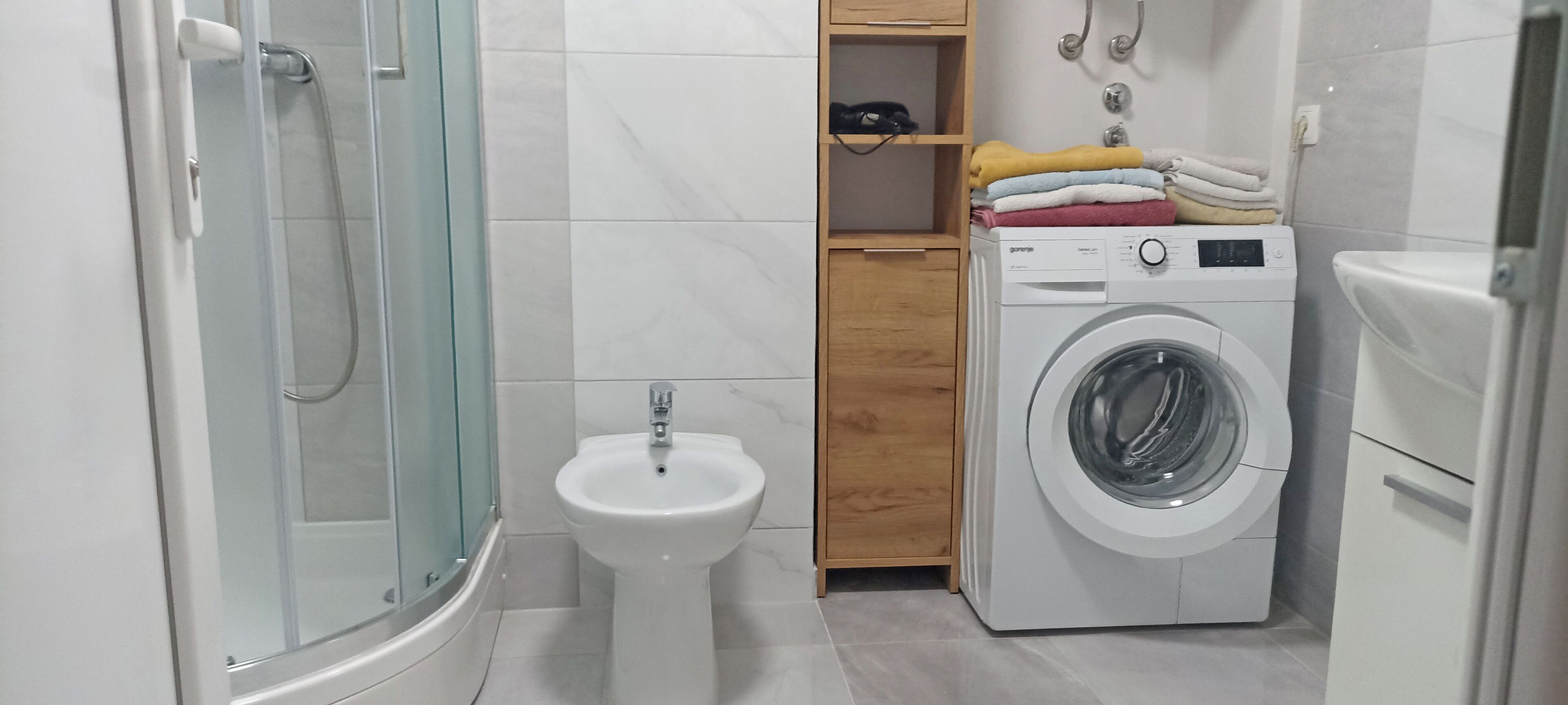 Un baño con ducha de vidrio, lavamanos sobre pedestal, lavadora y un armario de madera con toallas.