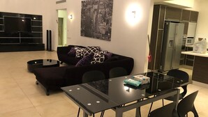 A modern living room with a black sofa, a glass dining table, and a kitchen area with a refrigerator and microwave. - Villa with Garden near Beach (Herzliya)