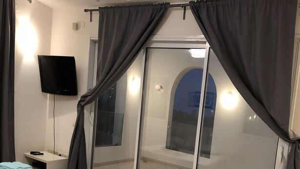 A hotel room with a bed, a bedside table with a remote control, a TV mounted on the wall, and a sliding glass door leading to an outdoor area. - Villa with Garden near Beach (Herzliya)