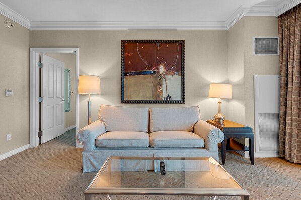 Habitación de hotel con un sofá color beige, una mesa de centro de vidrio, dos lámparas, un cuadro en la pared y una puerta que conduce a otra habitación.