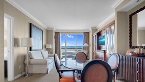 A hotel room with a dining table, chairs, a sofa, a TV, and a large window with a view of the beach.