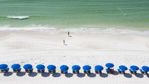 Ein Strand mit klarem türkisblauem Wasser, einem sandigen Ufer und mehreren blauen Sonnenschirmen und Liegestühlen.