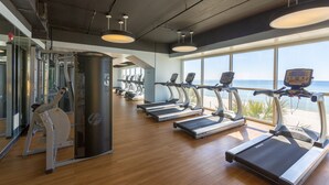 Ein Fitnessraum mit Laufbändern, Kraftgeräten und großen Fenstern mit Blick auf den Strand.