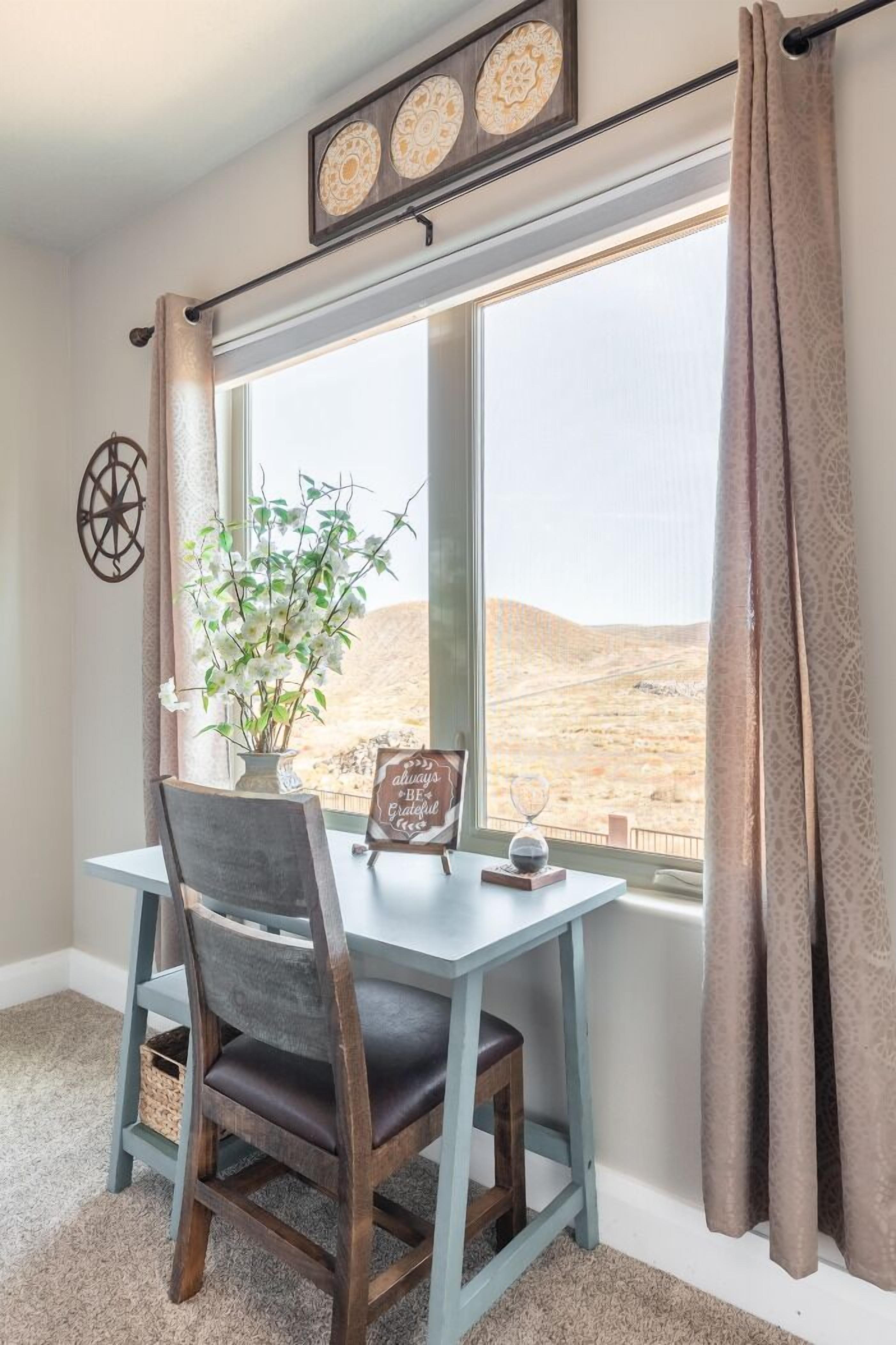 Un comedor con una mesa y sillas, una ventana que ofrece vistas a un paisaje desértico, un adorno en la pared y un cartel con un mensaje optimista.