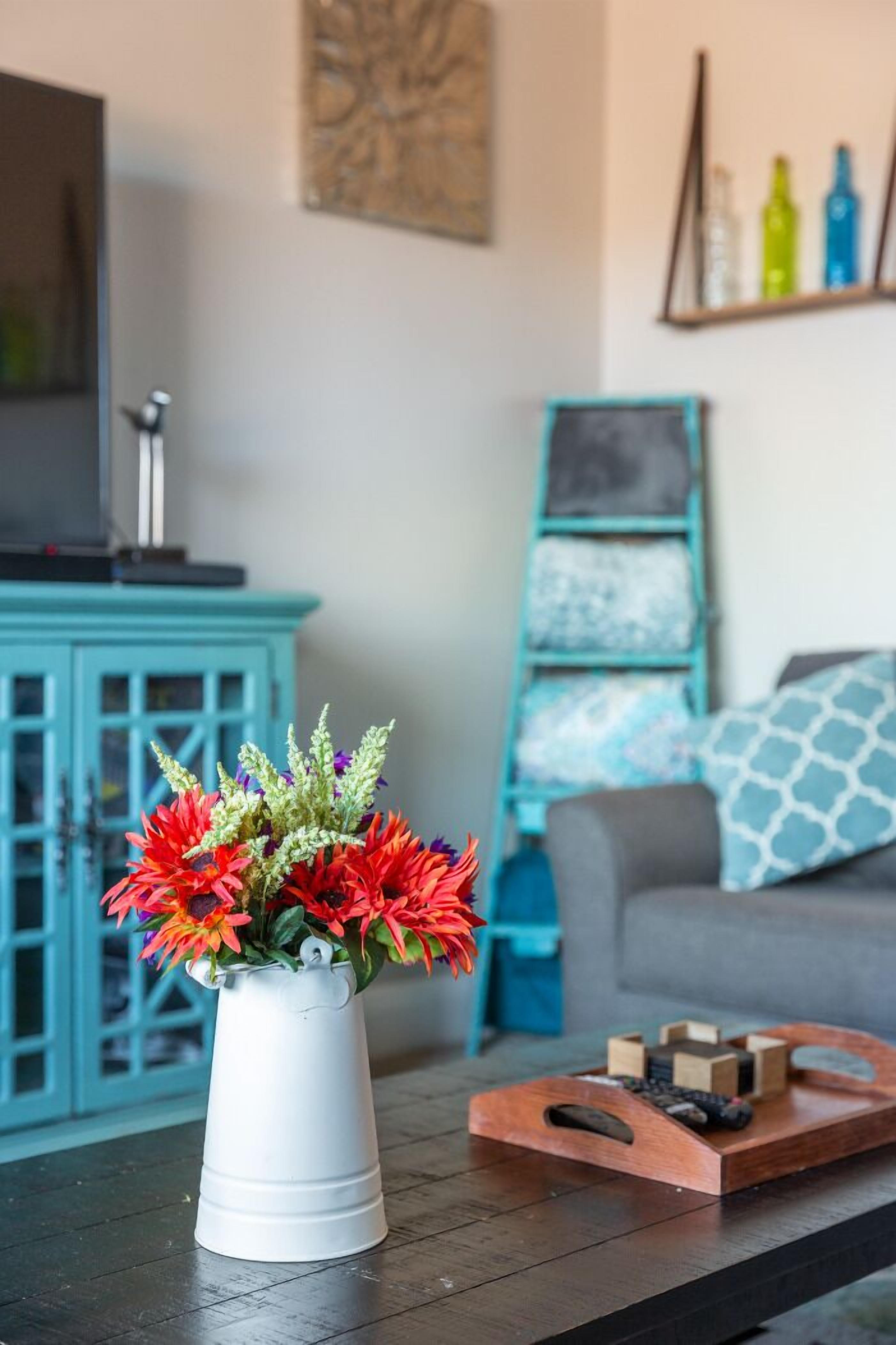 Un salón con un jarrón blanco lleno de flores coloridas sobre una mesa de centro de madera, un aparador azul verdoso, un sofá gris con cojines estampados y una estantería con botellas de colores.