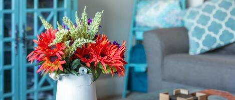 Un salón con un jarrón blanco lleno de flores coloridas sobre una mesa de centro de madera, un aparador azul verdoso, un sofá gris con cojines estampados y una estantería con botellas de colores.