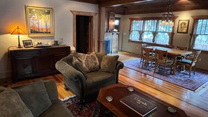 A cozy living room with a fireplace, a dining area with a wooden table and chairs, and a large window with curtains. - Historic Riverside Cottage ~ A Beautifully Restored 1908 Cottage! (Estes Park)