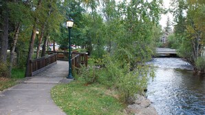 A concrete pathway alongside a river, bordered by trees and a wooden bridge. - Historic Riverside Cottage ~ A Beautifully Restored 1908 Cottage! (Estes Park)