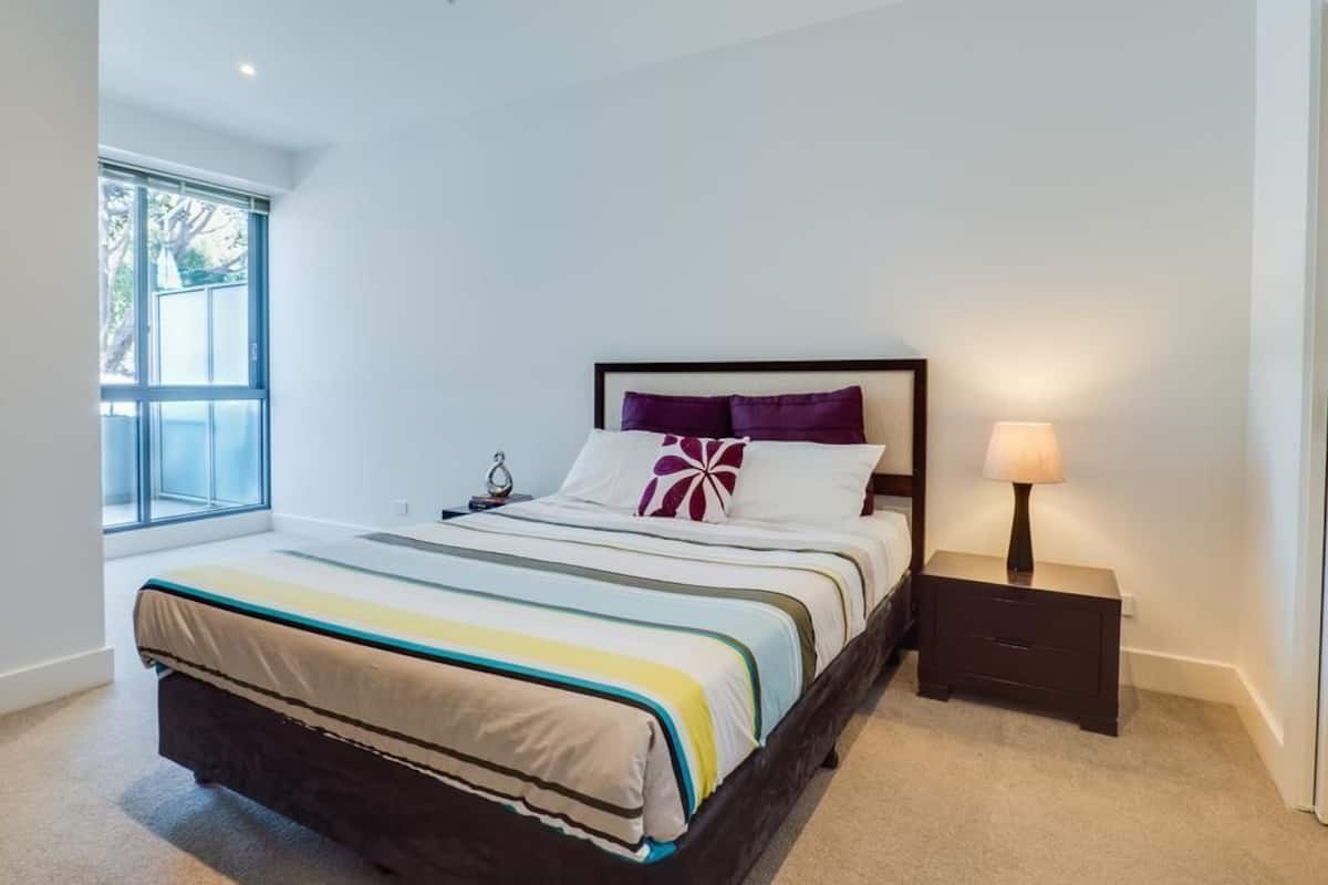 A bedroom with a large bed, a nightstand, a lamp, and a window with blinds.