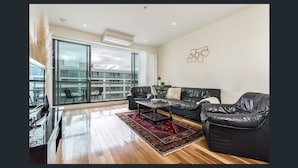 A modern living room with a black leather sofa, a glass sliding door leading to a balcony, and a large window offering a city view. - Waterfront By Gold Star Stays (Geelong)