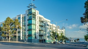 A modern multi-story building with a flag on top, located at a street corner with traffic lights and trees. - Waterfront By Gold Star Stays (Geelong)