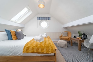 A bedroom with a bed, a round window, a skylight, a small table with a plant, and a chair.
