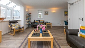 Een ruime woonkamer met een houten salontafel, een leren bank en een fauteuil met een gestreept kussen. Er is een boekenkast en een raam met uitzicht op het omliggende landschap.