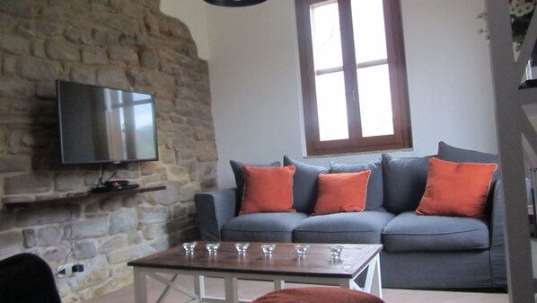 A living room with a stone wall, a grey sofa with orange cushions, a flat-screen TV mounted on the wall, and a small table with glasses.