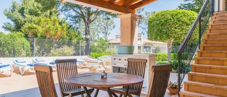 Une terrasse extérieure avec une table et des chaises en bois rondes, un barbecue et un escalier menant à un niveau supérieur.
