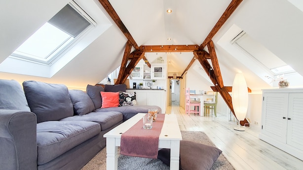 A loft-style room with a high ceiling, exposed wooden beams, a grey sofa, a white coffee table, and a kitchen area in the background. - Friendly Home - "Helles Dachstudio" - Cologne - Fair - Bonn - Phantasialand (Bornheim)