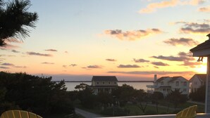 A deck with two yellow Adirondack chairs and a small table, overlooking a body of water and houses during sunset. - Two Crabby: Feels like home! Spectacular views, Beach Club Summer 2026! (Nags Head)