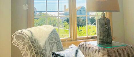A cozy room with a large window, a chair, a lamp, and a view of greenery and buildings.