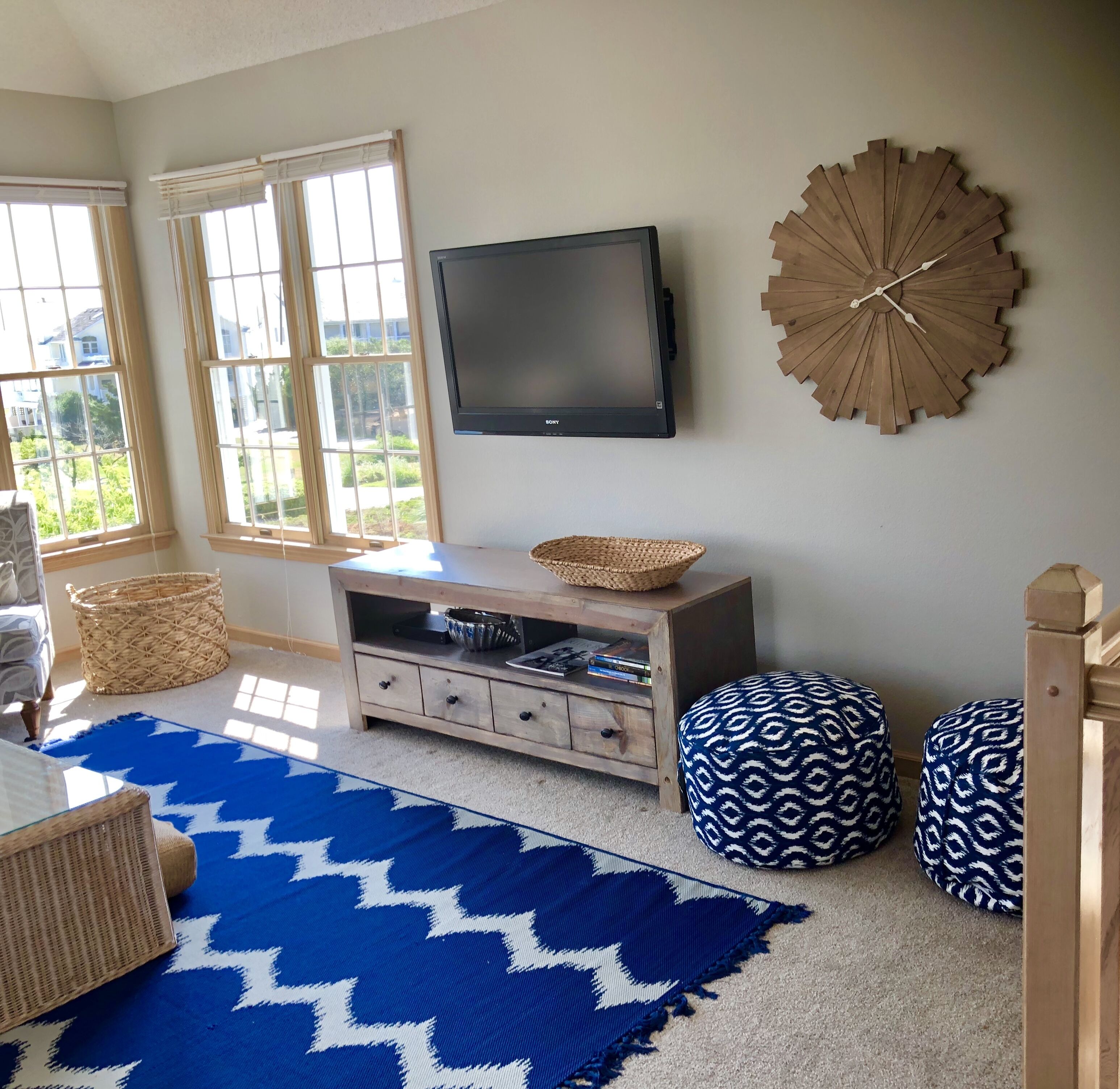 A room with a large window, a flat-screen TV, a wooden clock, and a blue patterned rug.