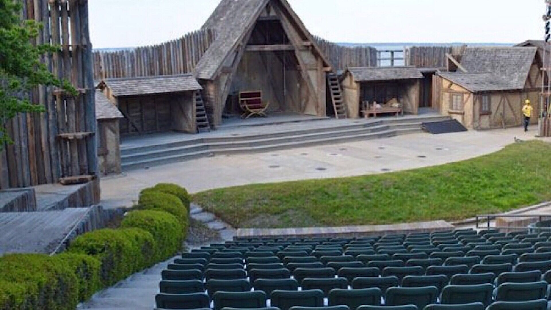 A rustic wooden stage with a backdrop of a large barn-like structure, surrounded by a grassy area and rows of green seats.