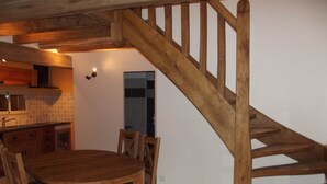Une table à manger en bois avec des chaises, un coin cuisine avec un pare-feu en carreaux, et un escalier en bois.