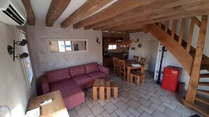 Un salon chaleureux avec un plafond en bois, un canapé rouge, une table à manger avec des chaises, et une cuisine en arrière-plan.