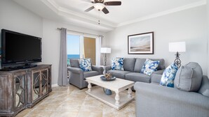 A living room with a flat-screen TV, grey sofas with blue and white patterned cushions, a wooden cabinet, and a view of the ocean through a large window. - Golf Cart ~ Beach Front ~ Balcony ~ Community Pool (Miramar Beach)