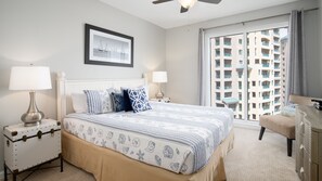A bedroom with a bed, two bedside tables, a ceiling fan, a window with a view of a building, and a framed picture on the wall. - Golf Cart ~ Beach Front ~ Balcony ~ Community Pool (Miramar Beach)