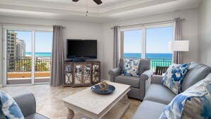 A spacious living room with a large flat-screen TV, grey sofa with blue patterned cushions, a wooden coffee table, and a view of the ocean through large windows. - Golf Cart ~ Beach Front ~ Balcony ~ Community Pool (Miramar Beach)