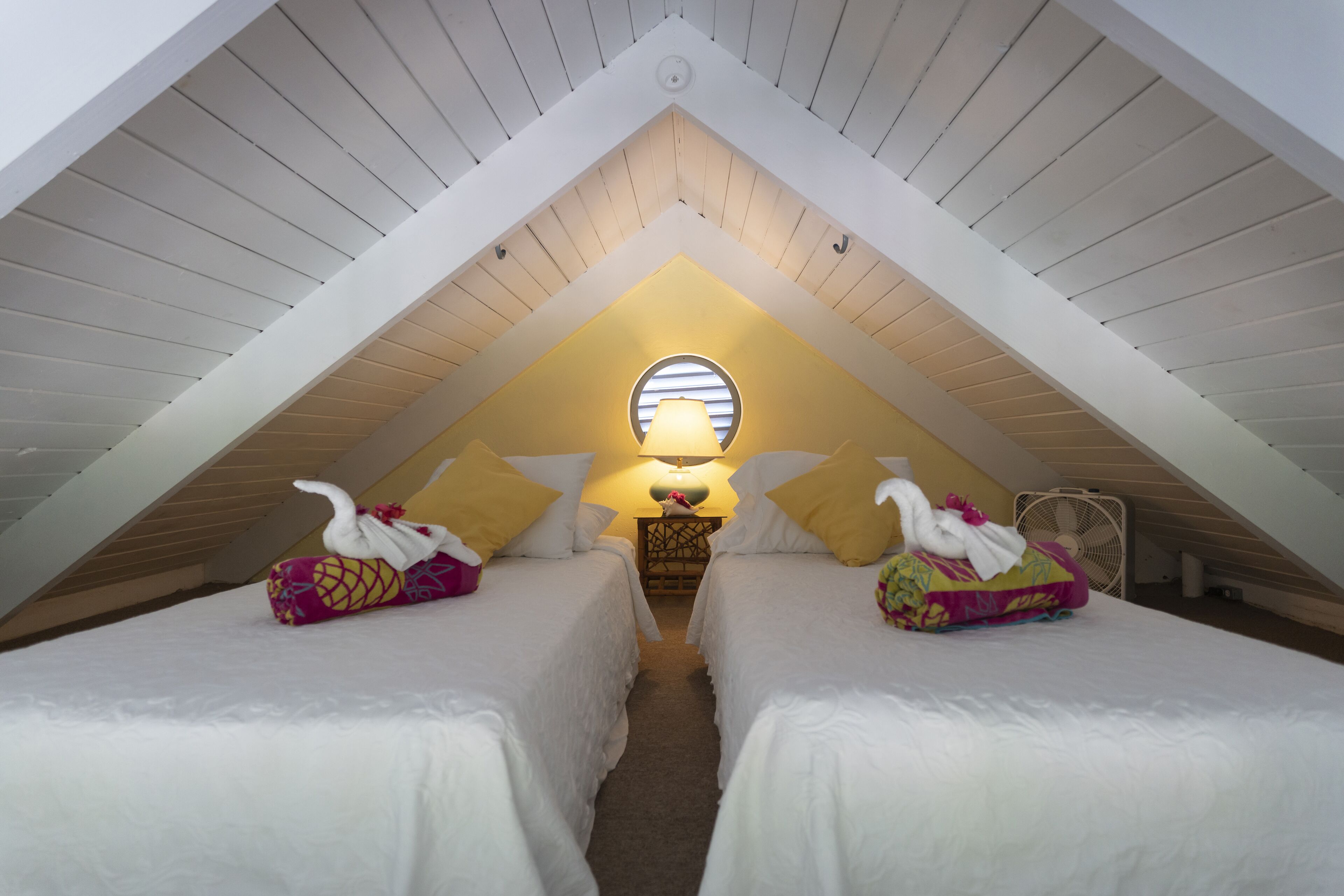 A cozy attic bedroom with two beds, a small table with a lamp, and a fan.
