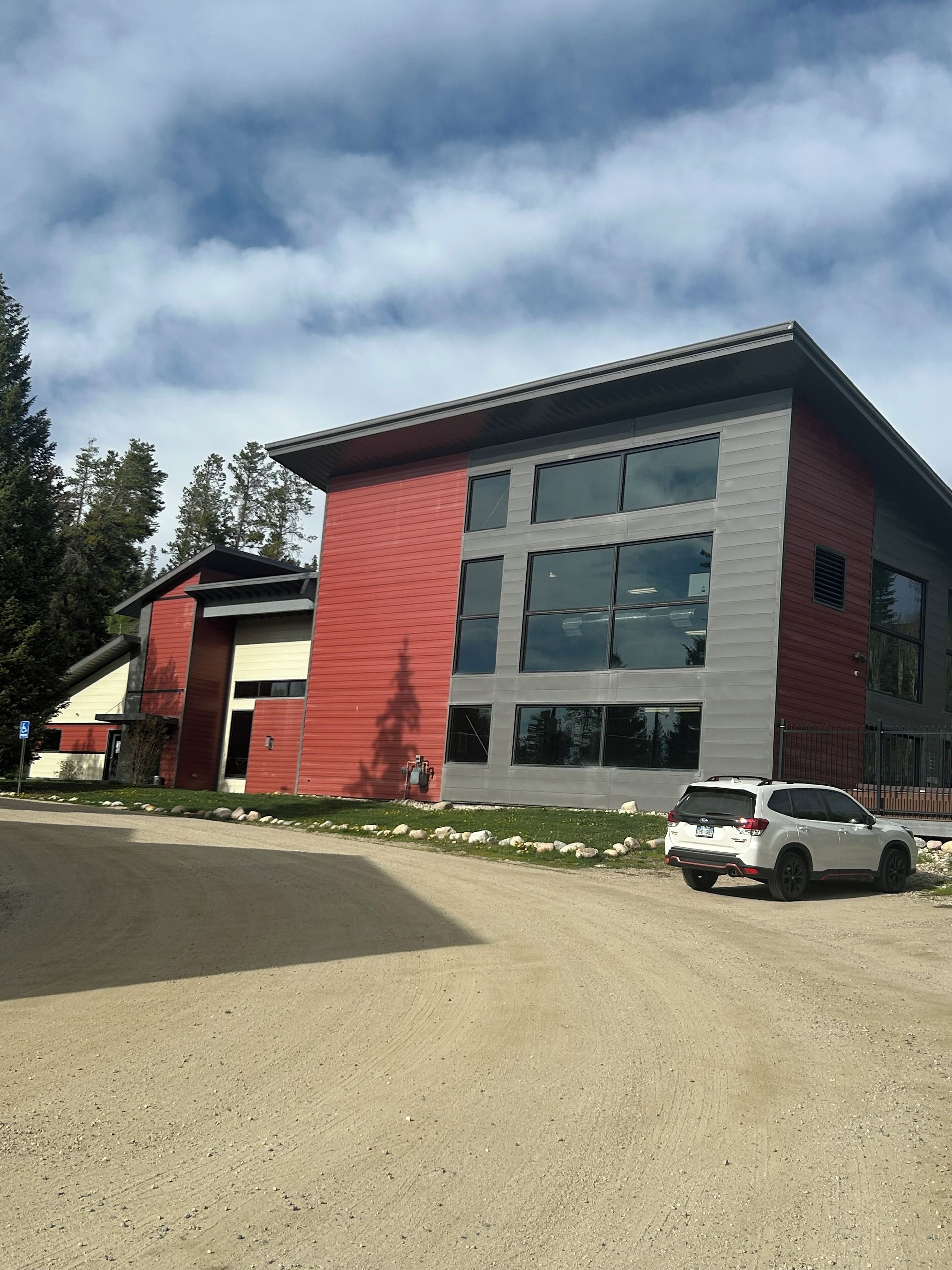 A modern building with a red and grey facade, large windows, and a parking area with a white SUV.